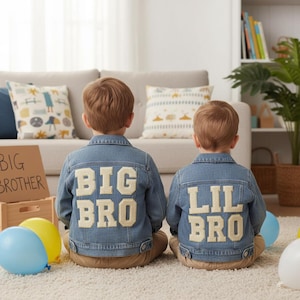 May include: Two children wearing denim jackets with the words "BIG BRO" and "LIL BRO" appliqued on the back. The jackets are light blue. The children are sitting on a white rug. Balloons and a sign that says "BIG BROTHER" are in the foreground.