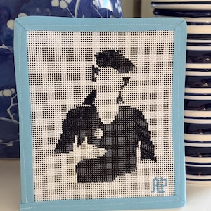May include: A square needlepoint art piece featuring a black and white silhouette of a person. The artwork is framed with a light blue fabric border and has the initials "AP" in the lower right corner. Decorative home decor.