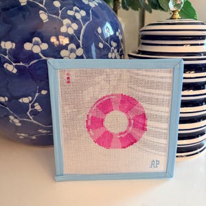 May include: A square needlepoint featuring a pink life preserver design on a white background, framed with a light blue border. The initials "AP" are in the lower right corner. Decorative blue and white vases are in the background.