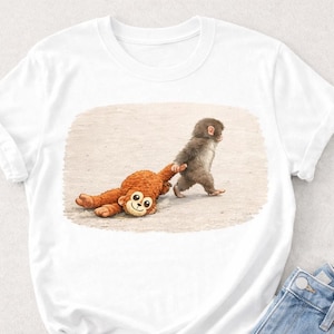 Pode incluir: Camiseta branca com uma ilustração de um macaco bebê puxando um macaco de pelúcia marrom. O brinquedo tem um rosto sorridente. O desenho é impresso em uma camiseta branca.