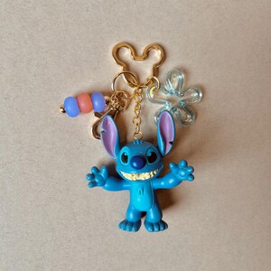 Pode incluir: Um chaveiro Stitch azul com um porta-chaves e berloques dourados. A figura Stitch tem orelhas grandes e est&aacute; em uma pose acolhedora. Berloques adicionais incluem uma flor transparente e uma corrente de contas com contas azuis, laranja e rosa.