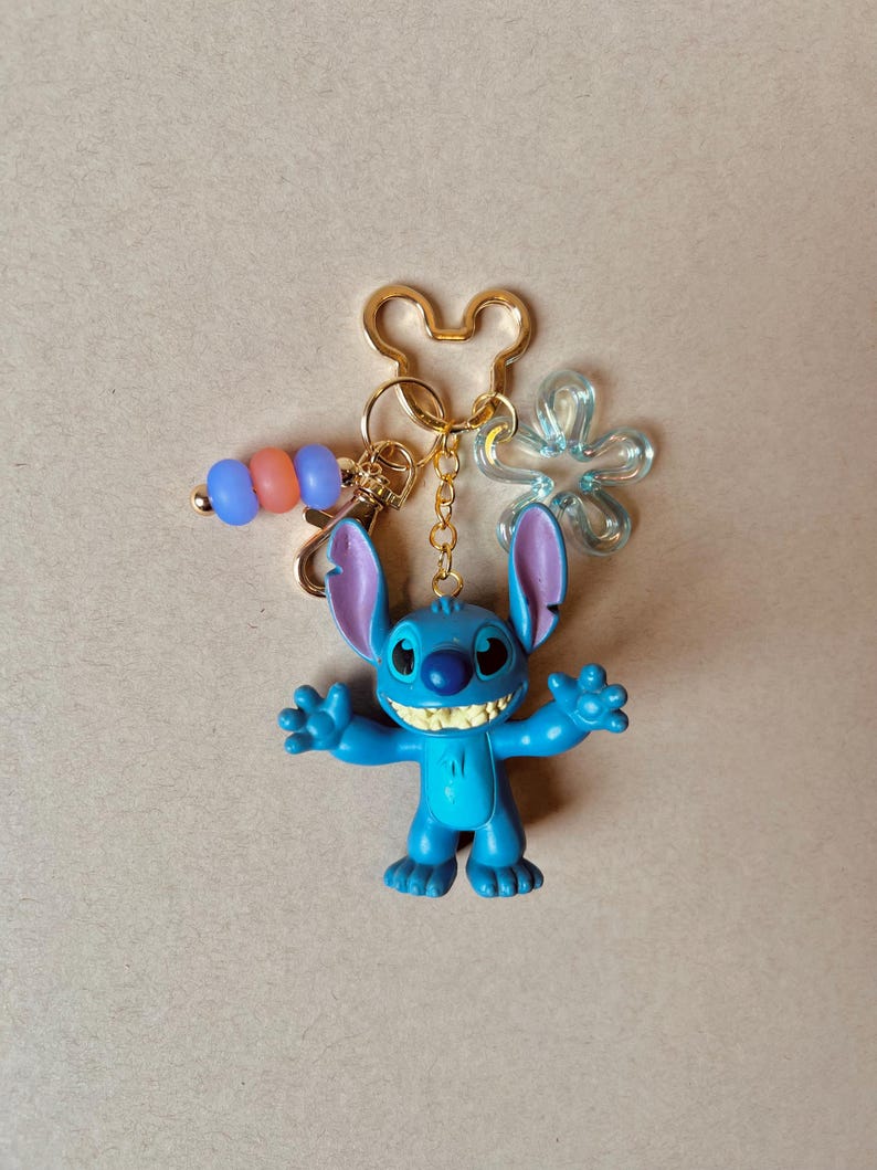 Pode incluir: Um chaveiro Stitch azul com um porta-chaves e berloques dourados. A figura Stitch tem orelhas grandes e est&aacute; em uma pose acolhedora. Berloques adicionais incluem uma flor transparente e uma corrente de contas com contas azuis, laranja e rosa.