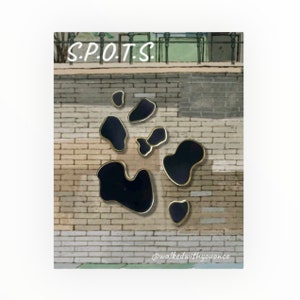 May include: A set of five black enamel pins in the shape of irregular spots, outlined in gold, against a brick wall background. The text "S.P.O.T.S." is visible at the top of the image.