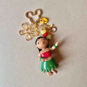 Pode incluir: Um chaveiro com uma figura de um personagem com top vermelho e saia verde, com um pingente dourado da cabe&ccedil;a do Mickey Mouse e uma flor. O chaveiro tamb&eacute;m inclui um mosquet&atilde;o e um grupo de contas coloridas.