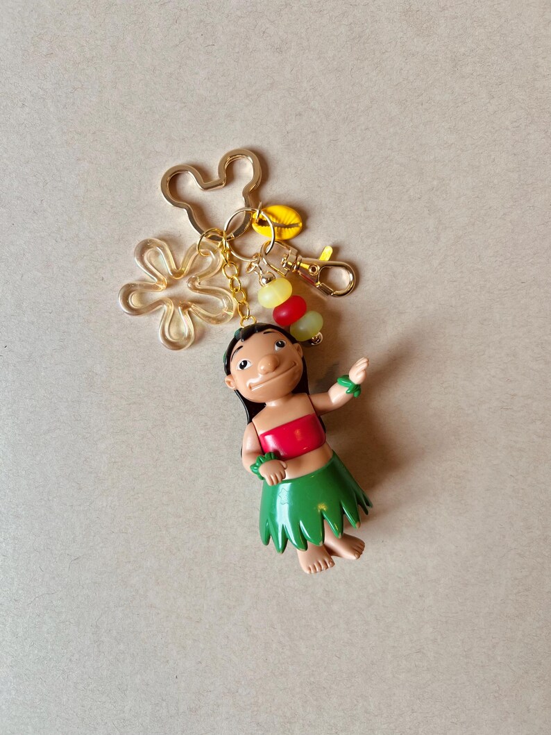 Pode incluir: Um chaveiro com uma figura de um personagem com top vermelho e saia verde, com um pingente dourado da cabe&ccedil;a do Mickey Mouse e uma flor. O chaveiro tamb&eacute;m inclui um mosquet&atilde;o e um grupo de contas coloridas.