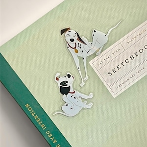 May include: Two Dalmatian dog stickers on a light green sketchbook. The stickers feature black and white spotted dogs with red collars. The sketchbook has a green spine with the text "VIVRE AVEC INTENTION" and a label that reads "SKETCHBOOK".