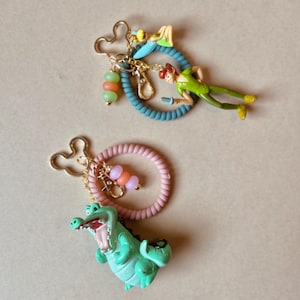 May include: Two keychains featuring cartoon characters. One keychain has a blue ring with a Peter Pan figure and a Tinkerbell figure. The other keychain has a pink ring with a crocodile figure. Both keychains have gold-colored Mickey Mouse-shaped clasps and beaded accents.