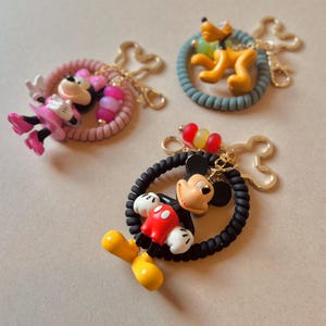 May include: Three character keychains featuring Mickey Mouse, Minnie Mouse, and Pluto. Each keychain has a gold-colored clasp and a coiled ring in various colors. Mickey Mouse wears red shorts and yellow shoes. Minnie Mouse wears a pink dress and shoes.
