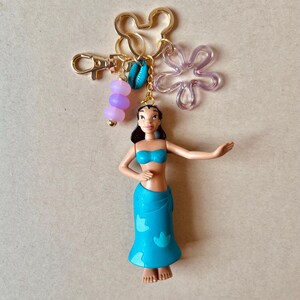 Pode incluir: Um porta-chaves dourado com uma pequena figura de uma mulher com top e saia de biqu&iacute;ni azuis. O porta-chaves inclui um pingente em forma de Mickey Mouse, uma flor transparente, contas roxas e uma concha.