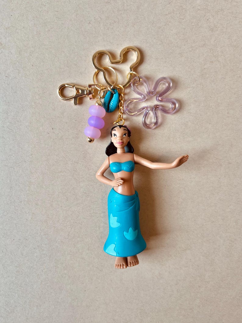 Pode incluir: Um porta-chaves dourado com uma pequena figura de uma mulher com top e saia de biqu&iacute;ni azuis. O porta-chaves inclui um pingente em forma de Mickey Mouse, uma flor transparente, contas roxas e uma concha.