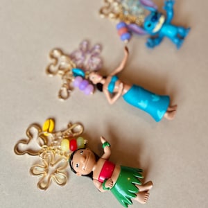 Pode incluir: Tr&ecirc;s figuras de personagens coloridos, incluindo um alien azul, uma menina com top vermelho e saia verde, e uma mulher com roupa azul. Cada figura &eacute; acompanhada por um pingente dourado com contas coloridas. O fundo &eacute; bege neutro.