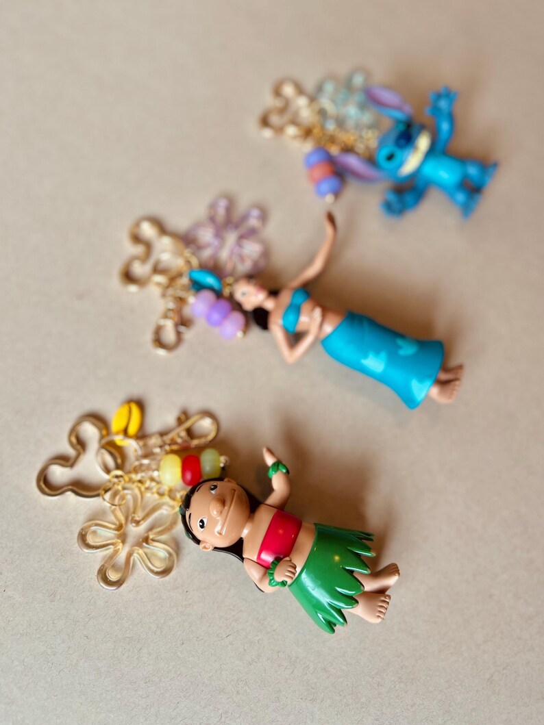 Pode incluir: Tr&ecirc;s figuras de personagens coloridos, incluindo um alien azul, uma menina com top vermelho e saia verde, e uma mulher com roupa azul. Cada figura &eacute; acompanhada por um pingente dourado com contas coloridas. O fundo &eacute; bege neutro.