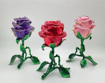 May include: Three decorative rose sculptures in purple, red, and pink. Each rose has a detailed, jewel-encrusted bloom atop a green stem with leaves. The stems are supported by a three-pronged base.
