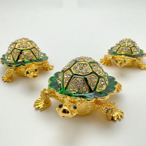 Puede incluir: Tres figuras decorativas de tortugas. Cada tortuga tiene un cuerpo y patas doradas, un caparazón verde con borde festoneado y está incrustada con cristales transparentes. Las tortugas están dispuestas sobre una superficie blanca.
