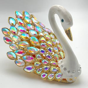 May include: A decorative swan figurine with a white body, gold accents, and iridescent, oval-shaped jewels forming the tail. The swan has a gold beak and a small blue eye. The base is gold and the body is decorated with small, clear crystals.