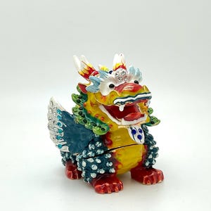May include: A colourful, detailed dragon figurine with a red, yellow and green body, blue wings, and a white and blue face. The dragon is decorated with rhinestones and has a hinged jaw.