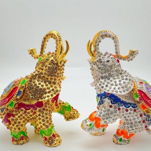 May include: Two bejeweled elephant figurines. One is gold-toned, the other silver-toned, both adorned with colorful enamel accents. The elephants have raised trunks and are covered in small, sparkling crystals. The figurines are decorative and ornate.