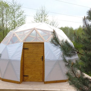 May include: A white geodesic dome tent with a wooden door and a wooden frame. The tent is surrounded by trees.