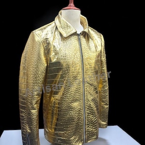 May include: A gold-colored jacket with a textured, crocodile-like pattern. The jacket has a zipper closure, a collar, and a zipper detail on the sleeve. The jacket is displayed on a mannequin against a black background.