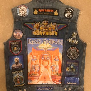 Puede incluir: Un chaleco vaquero azul con varios parches de la banda Iron Maiden, incluyendo la portada del álbum "Powerslave", un parche de la gira mundial "World Slavery Tour" y varios parches de Eddie the Head.
