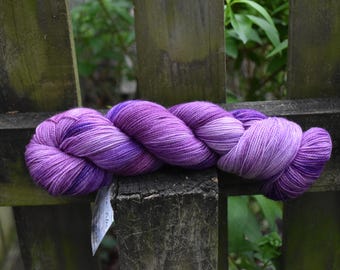Può includere: Un gomitolo di filato viola, con varie sfumature di viola e lavanda, è appoggiato su una staccionata di legno usurata. Il filato è ben avvolto, mostrando la sua consistenza e le variazioni di colore. Una piccola etichetta è attaccata al filato.