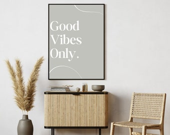 May include: A framed print with a gray background and white text that reads "Good Vibes Only." The print is hanging on a white wall above a wooden cabinet with a black metal frame. A woven chair is in the foreground.