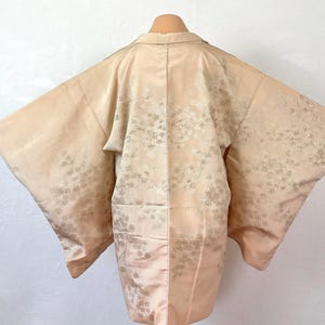 以下が含まれることがあります： 淡いピーチカラーの羽織で、生地全体に繊細な同系色の花柄が施されています。ゆったりとした袖としっかりとした襟が特徴で、表面全体にほのかに光沢のある仕上がりが見られます。