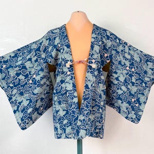 以下が含まれることがあります： 白と水色の花柄が全体に施された、袖の広い青いジャケット。前開きデザインで、胸元には小さな装飾用の紐留めが付いており、生地全体に植物のプリントが繰り返されています。