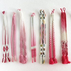 Può includere: Una collezione di dieci cordoncini decorativi in varie tonalità di rosa, bianco e rosso. I cordoncini presentano intricati motivi intrecciati, nappe e nodi decorativi. Alcuni cordoncini hanno un effetto sfumato, che passa dal rosa chiaro allo scuro. I cordoncini sono disposti su uno sfondo bianco.