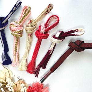 May include: An assortment of six decorative Japanese obi jime cords in various colors. The cords are knotted and feature tassels. Colors include navy blue, pink, gold, cream, red, and burgundy. The cords are arranged on a white background.