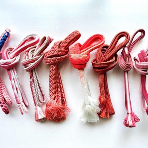 May include: A collection of eight decorative cords in shades of pink, red, and white. Each cord is knotted at the top and features a tassel at the bottom. The cords vary in design, with some having striped patterns and others solid colors.