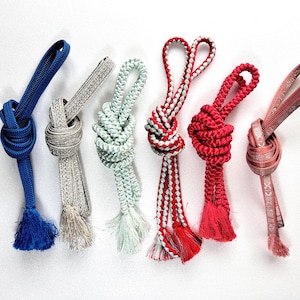 May include: Six decorative cords in various colors, including blue, gray, mint green, red and pink. Each cord is knotted and has a tassel at the end. The cords are arranged on a white background.