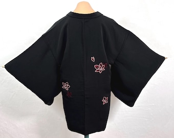 May include: Black kimono-style jacket with wide sleeves. The back of the jacket features embroidered red and white floral designs. The jacket is displayed on a mannequin, showcasing its shape and design. The garment is made of a smooth, dark fabric.