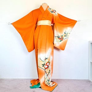 May include: An orange kimono with a white obi belt. The kimono features floral designs in red, yellow, and green. The fabric has a gradient effect, transitioning from orange to white. The sleeves are long and wide.