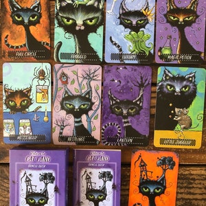 May include: A purple and white oracle deck with a black cat illustration on the front. The deck is titled "Return to Cat Land Oracle Deck" and is by Joanna Nelson. The cards are spread out on a wooden surface, showing various illustrations of black cats with different backgrounds.