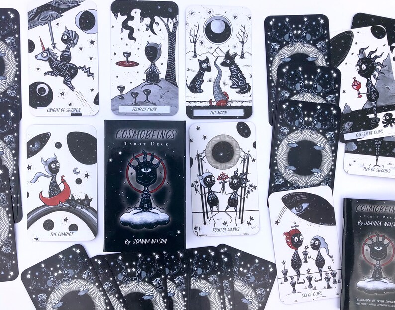 May include: A black and white tarot deck with a dark, starry background. The cards feature illustrations of various characters and symbols. The deck is titled "Cosmobeings Tarot Deck" and is by Joanna Nelson.