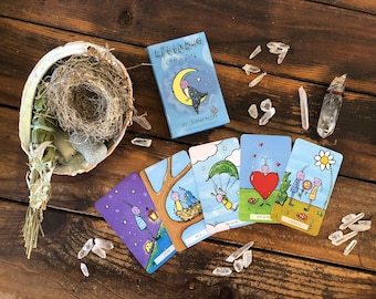 May include: A set of colorful oracle cards with illustrations of bugs and nature scenes. The cards are laid out on a wooden surface with a nest, crystals, and a sage bundle. The box for the cards is visible in the upper right corner and says "Little Bug Oracle Deck" by Jeannine Nielsen.