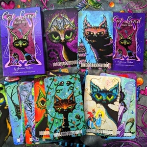May include: A set of Cat Land Oracle Deck cards featuring colorful illustrations of cats with whimsical designs. The cards include titles like "Inner Monster", "Wild Thang", and "Create".