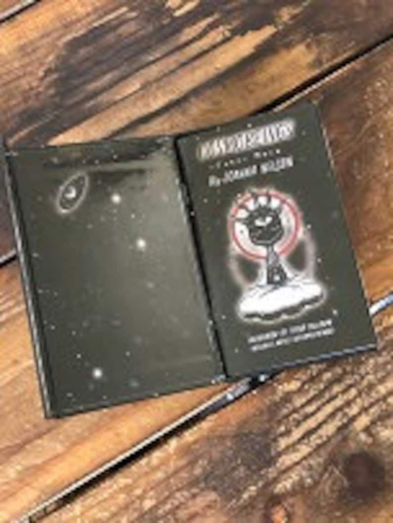 May include: A black hardcover book with a white illustration of a black cat on a cloud with a crescent moon and stars in the background. The book title is "Don't Worry, It's Just a Dream" by Joanna Hollen. The book cover is illustrated with a black cat with a white eye and a white crescent moon.