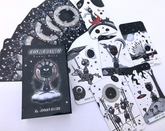 Puede incluir: Una caja negra con el texto "COSMOBEINGS TAROT DECK" y "By JOANNA NELSON". La caja está abierta y muestra una baraja de tarot con ilustraciones en blanco y negro de criaturas fantásticas y cuerpos celestes. Las cartas están extendidas sobre una superficie blanca.