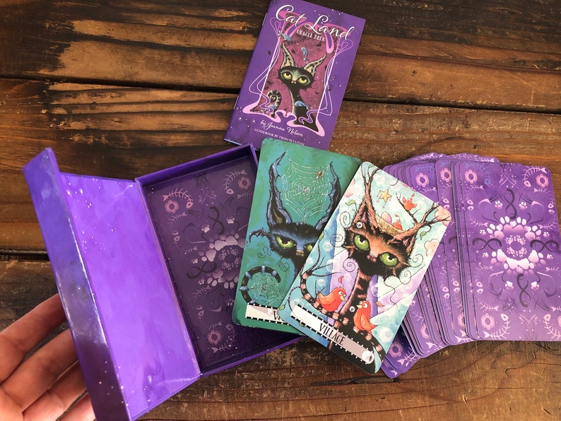 May include: A purple box with a deck of Cat Land Oracle Cards. The box is open and shows a purple interior with a floral pattern. Three cards are pulled out of the deck, showing colorful illustrations of cats with whimsical details. The cards are labeled "Village" and "The Web".