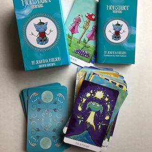 Może przedstawiać: Niebieskie i białe pudełko talii kart tarota z tytułem "Monstarot Tarot Deck" i tekstem "By Joanna Nelson Second Edition". Pudełko znajduje się obok niebieskiej i białej karty tarota z tekstem "The Luminary" i zieloną postacią potwora z ż&oacute;łtą aureolą.