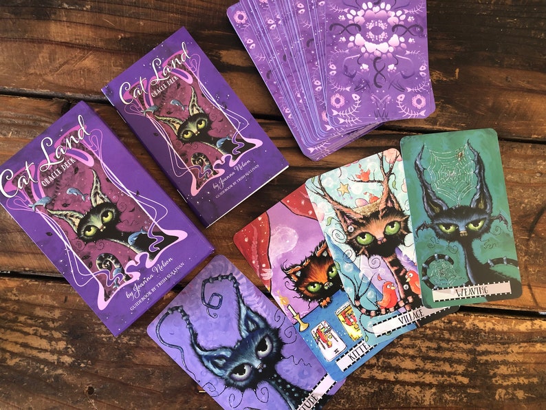 May include: A purple and white oracle deck called "Cat Land" by Joanna Nelson. The deck features illustrations of cats with whimsical and slightly spooky designs. The cards are laid out on a wooden surface.