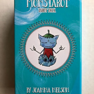 Może przedstawiać: Turkusowe pudełko z tekstem "Monstarot Tarot Deck" i "By Joanna Nelson Second Edition". Pudełko zawiera białe koło z kresk&oacute;wkową ilustracją niebieskiego stworzenia przypominającego kota z zielonym liściem na głowie, siedzącego w medytacyjnej pozie.
