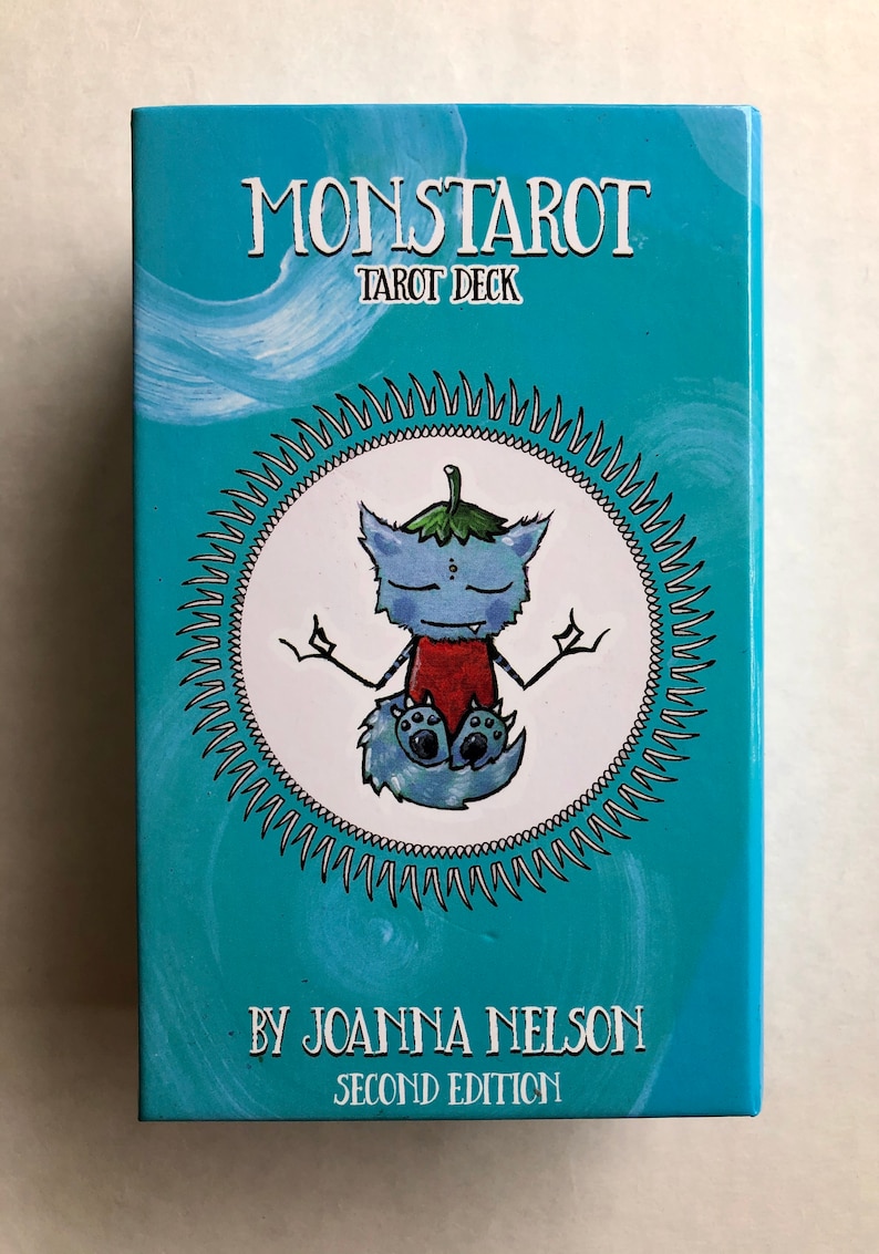 Może przedstawiać: Turkusowe pudełko z tekstem "Monstarot Tarot Deck" i "By Joanna Nelson Second Edition". Pudełko zawiera białe koło z kresk&oacute;wkową ilustracją niebieskiego stworzenia przypominającego kota z zielonym liściem na głowie, siedzącego w medytacyjnej pozie.