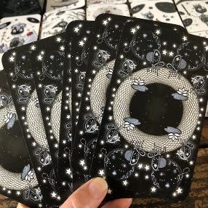 May include: A set of black tarot cards with a white star pattern and a silver snake design. The cards feature a black and white illustration of a snake with a crown on its head.