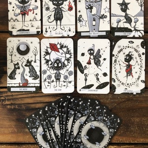 May include: A set of tarot cards with black and white illustrations of various characters and symbols. The cards are laid out in a spread on a wooden surface. The cards include Temperance, The Devil, The Tower, The Star, The Moon, The Sun, Judgement, and The World.