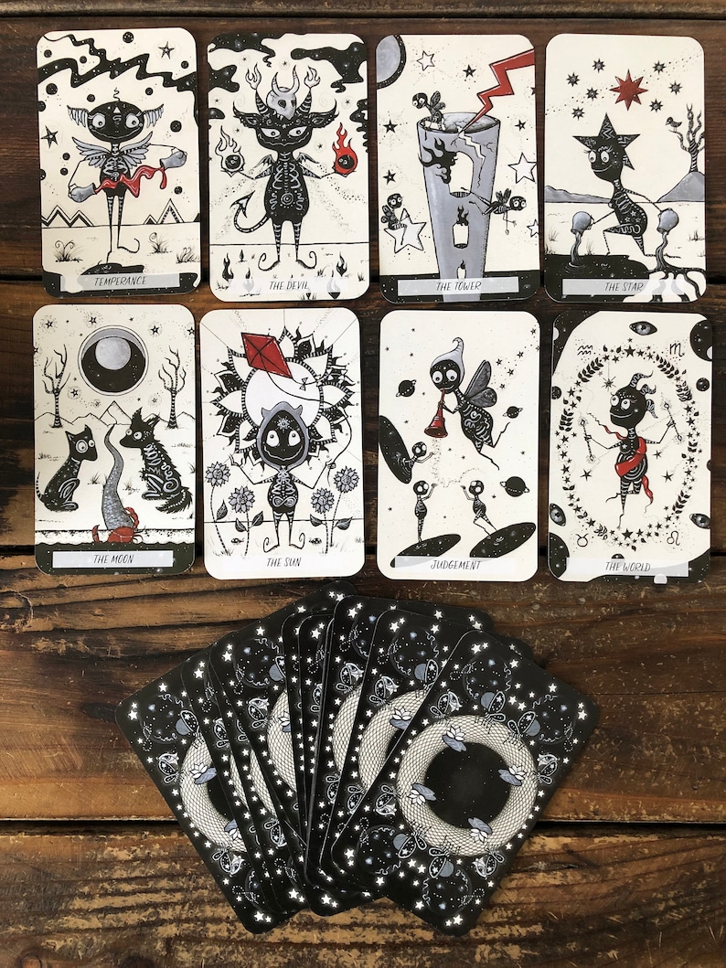 May include: A set of tarot cards with black and white illustrations of various characters and symbols. The cards are laid out in a spread on a wooden surface. The cards include Temperance, The Devil, The Tower, The Star, The Moon, The Sun, Judgement, and The World.