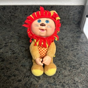 Może przedstawiać: Pluszowy lew z czerwoną grzywą i żółtym ciałem w kropki. Lew ma uśmiechniętą twarz z niebieskimi oczami i brązowym nosem.