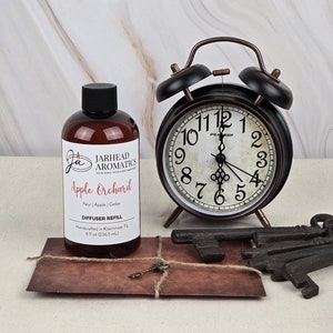 May include: A brown glass bottle with a label that reads "Jarhead Aromatics Apple Orchard Pear | Apple | Cedar Diffuser Refill 8 fl oz (236.5 mL)". The bottle is sitting on a brown surface next to a vintage alarm clock and several antique keys.